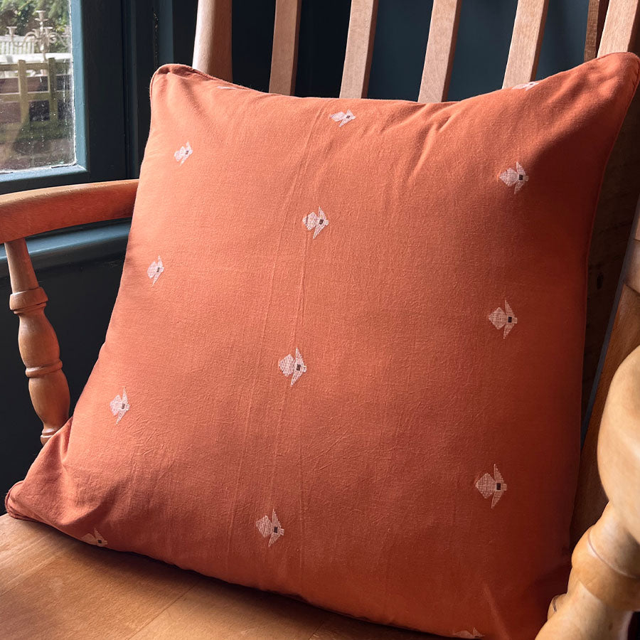 Terracotta Jamdani cushion cover with white motifs on a wooden chair by a window.