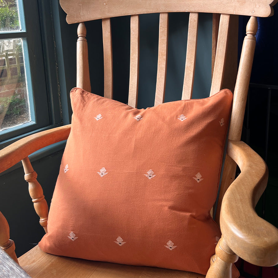 Terracotta Jamdani cushion cover with white motifs on a wooden chair by a window.