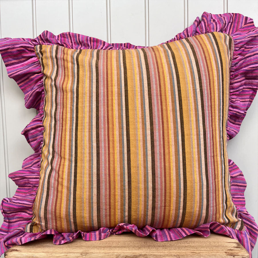 Striped yellow pillow with pink ruffled edges on a wooden surface against a white panelled wall.