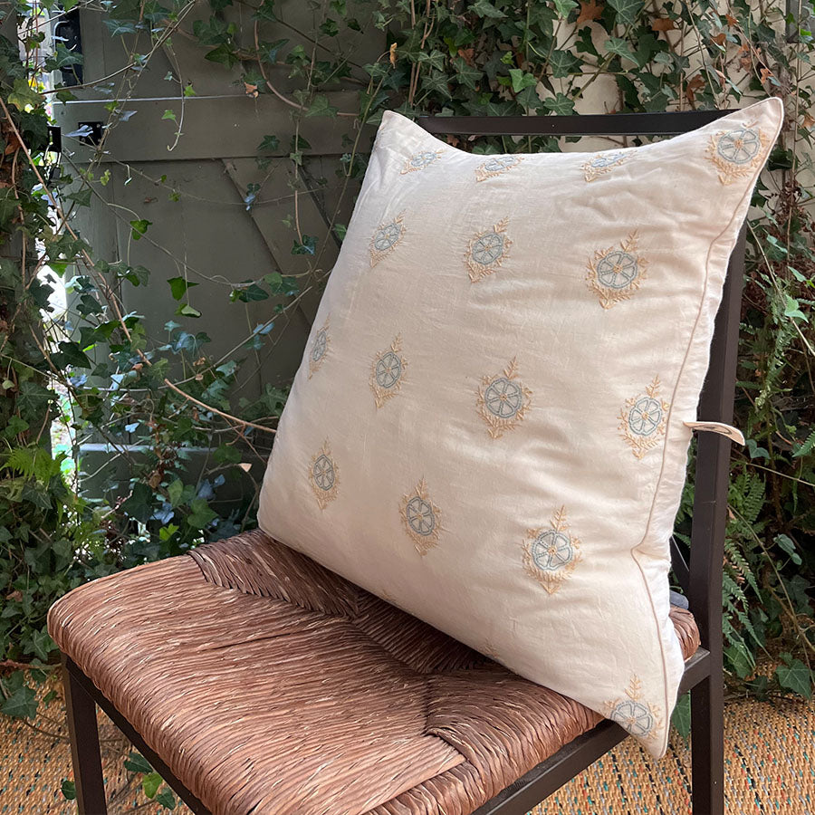 Bela Chikankari cushion cover with intricate blue and light beige motifs, placed on a chair outdoors.