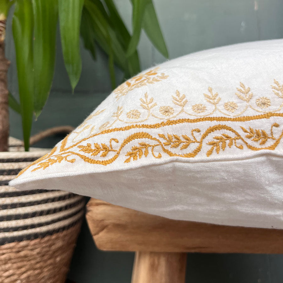 White cushion cover with gold embroidery on a wooden stool with a plant in the background
