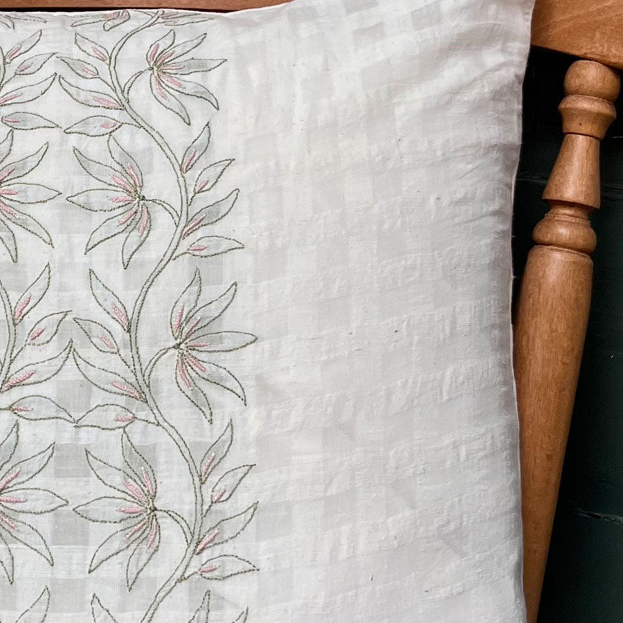 Close-up of chikankari cushion cover with lotus bloom embroidery placed on a wooden chair against a grey wall.